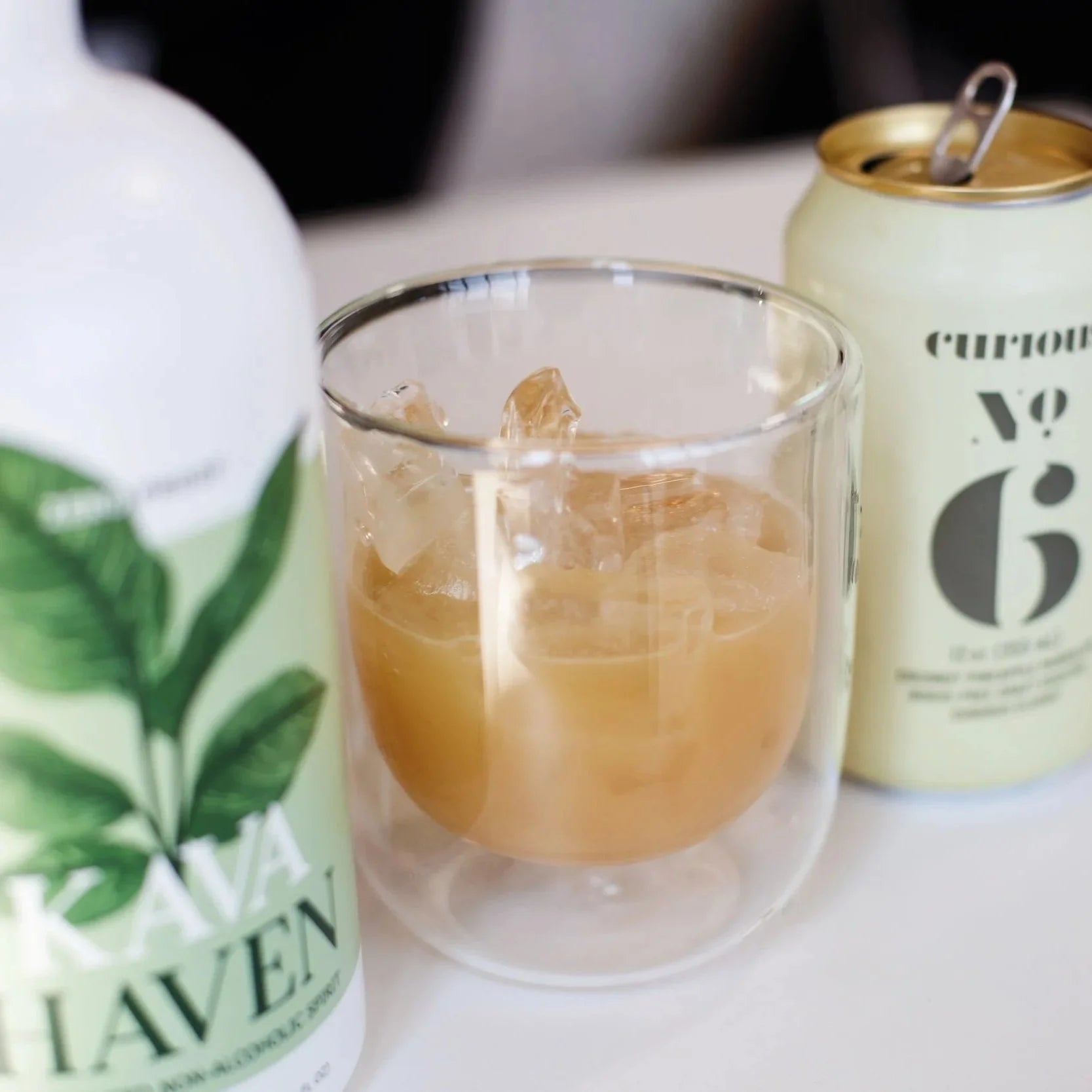 A Kava Haven bottle and Curious No. 6 can beside a glass of golden non-alcoholic cocktail over ice