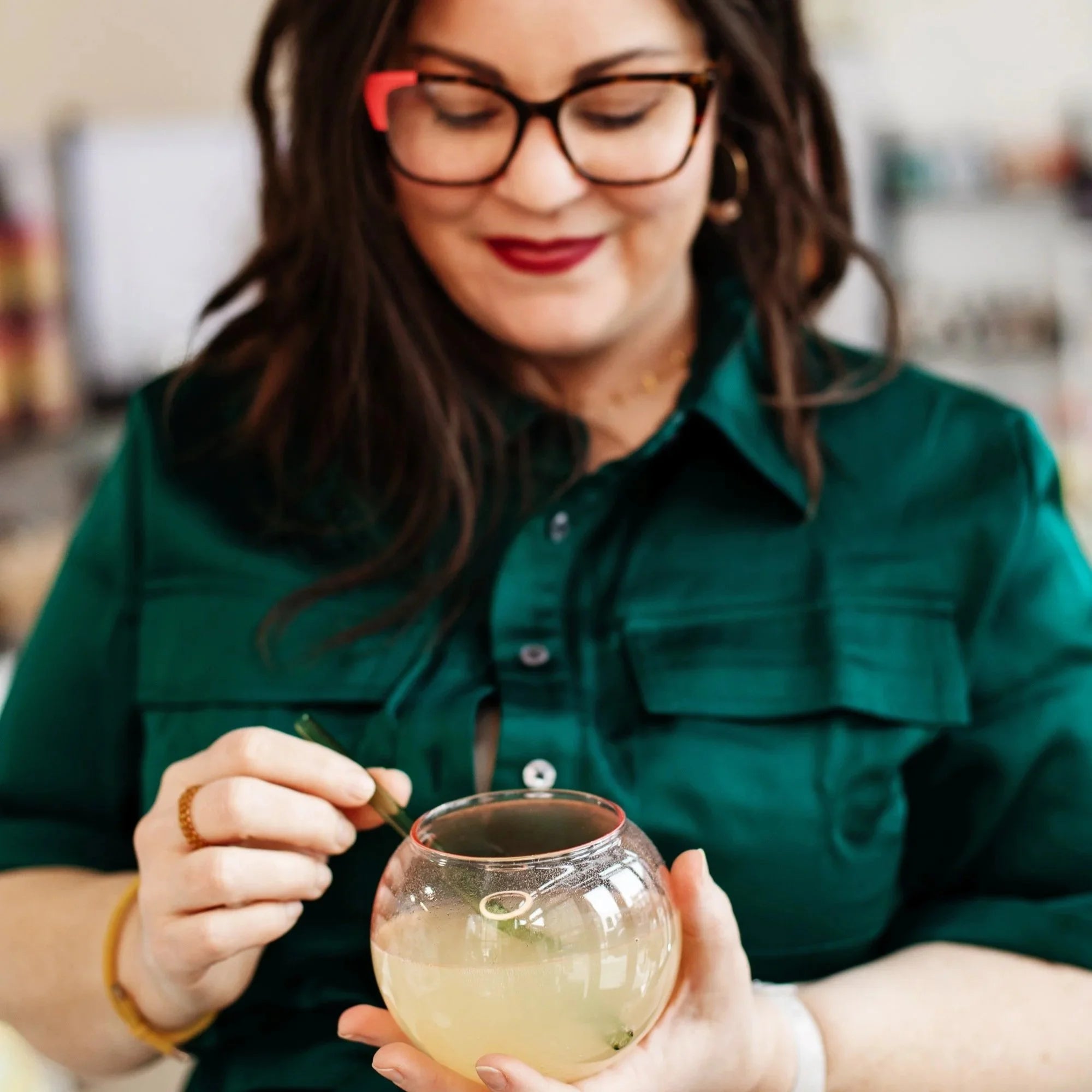 A person in a green shirt stirring a pale, herb-garnished non-alcoholic cocktail in a rounded glass