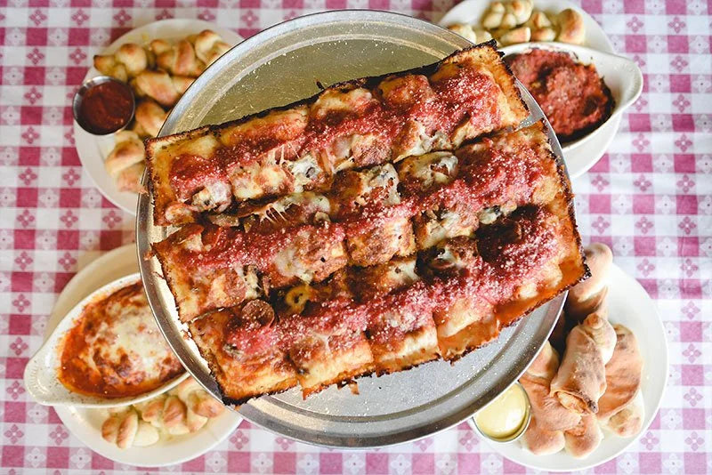 Detroit-style square pizza on a metal tray surrounded by sides of garlic knots and dips on a checked tablecloth